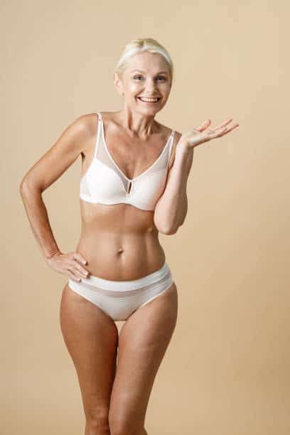Cheerful mature woman in underwear with fit body smiling at camera and holding hand on her waist, standing isolated over beige background. Beauty, Self acceptance concept