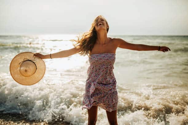 Mujer joven juguetona en vestido y sombrero disfrutando del viento y las olas en la playa